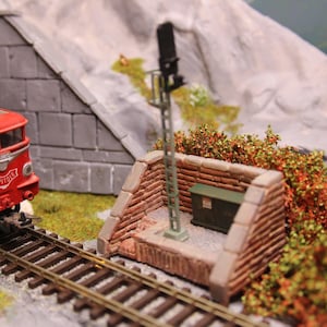 May include: A red and white model train engine with the word "CAPITOL" on the side, pulling into a tunnel. The train is on a model railway track with a small brick wall and a signal light on the right side of the track.