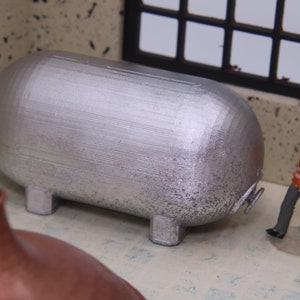 May include: A miniature silver industrial tank with a textured surface, resting on four small legs. A small figure of a worker in an orange vest and blue helmet stands nearby. The scene is set in a miniature factory or industrial environment.