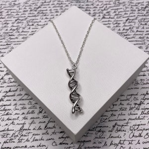 May include: Silver DNA helix pendant necklace. The pendant, a detailed DNA double helix, hangs from a delicate chain. The necklace is presented on a white surface, set against a backdrop of handwritten script.