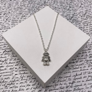 May include: A silver robot pendant necklace. The pendant, with a detailed design, hangs from a fine chain. The necklace is presented on a white surface, set against a backdrop of handwritten script. The robot pendant is a charming accessory.