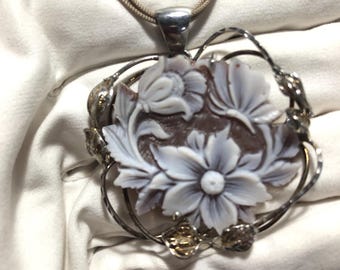 May include: A floral cameo pendant necklace. The pendant features a carved flower design in shades of gray and brown, set within a silver-toned frame. The necklace has a delicate, woven cord.