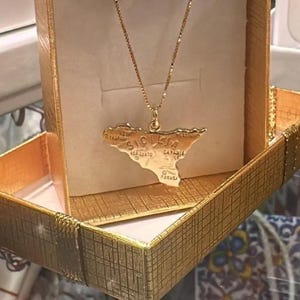 Può includere: Una collana dorata con un ciondolo a forma dell'isola di Sicilia. Il ciondolo reca l'iscrizione "Sicilia" e altri nomi di luoghi. La collana è presentata in un cofanetto regalo dorato.