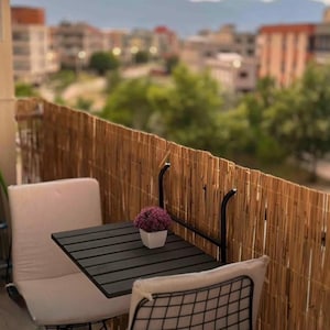 May include: A black metal table with a black wooden top sits on a balcony with a bamboo fence. Two chairs, one with a white cushion and one with a black mesh back, are positioned around the table. A small potted plant with purple flowers sits on the table.