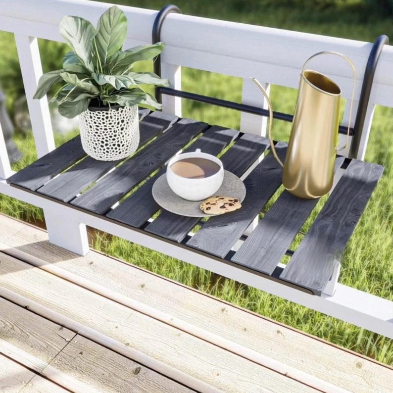 May include: A black wooden table with a metal frame attached to a white railing. The table has a cup of coffee and a cookie on a plate. There is a potted plant and a gold watering can on the table.