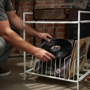 May include: White metal record storage rack with a turntable on top. A person is holding a black vinyl record. The rack holds several vinyl record albums. The turntable is brown with a gold mesh speaker.
