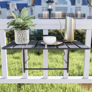 May include: A black metal folding table mounted on a white railing. The table has a wooden top and is holding a white cup, a plate, and a watering can.