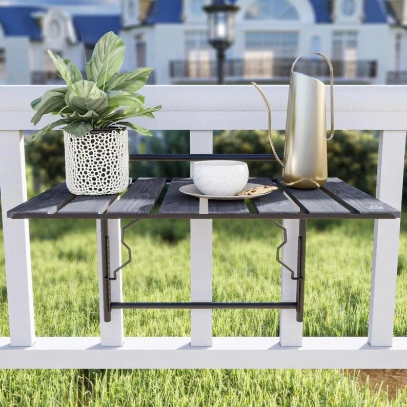 May include: A black metal folding table mounted on a white railing. The table has a wooden top and is holding a white cup, a plate, and a watering can.