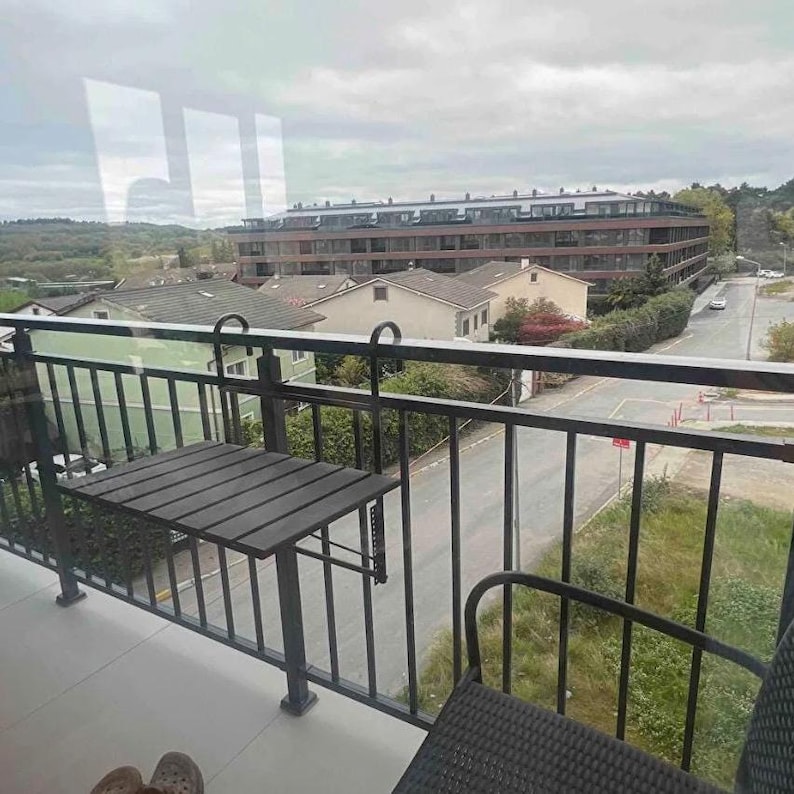 May include: A black metal folding table with a wooden top is mounted to a black metal railing on a balcony. The balcony overlooks a city street with buildings in the background.