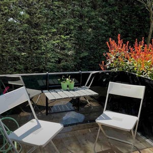May include: Two white folding chairs and a small wooden table with two potted plants on a balcony with a black railing and a green hedge in the background.