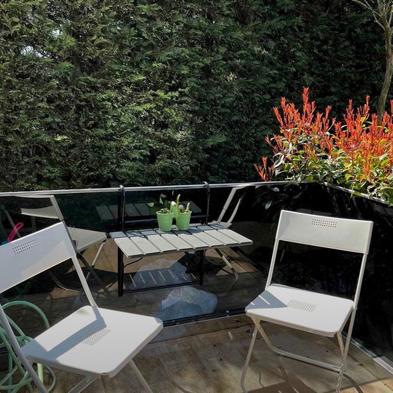 May include: Two white folding chairs and a small wooden table with two potted plants on a balcony with a black railing and a green hedge in the background.