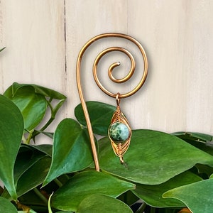 May include: A green stone pendant hangs from a copper wire plant stake with a spiral design. The stake is in a black pot with a green plant.