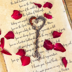 May include: A wire-wrapped heart-shaped pendant featuring small red beads, resting on an open book titled Invocation to Hecate. The page is decorated with scattered red rose petals and a black-and-white illustration of a figure holding a torch.