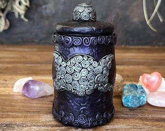 May include: A cylindrical dark purple jar with a removable lid, featuring a silver-toned triple moon symbol on the front. The surface is decorated with embossed spiral patterns and floral motifs, with a small spiral knob on the center of the lid.