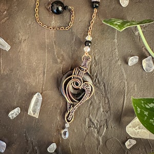 May include: A wire-wrapped pendant featuring a circular black stone encased in copper wire, shaped into a heart design. The pendant hangs from a copper chain adorned with black beads, clear faceted crystals, and a small clear teardrop-shaped charm.