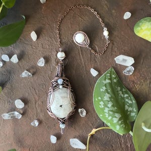 May include: A wire-wrapped pendant featuring a large, oval-shaped white moonstone encased in copper wire, accented with smaller white beads. The pendant hangs from a copper chain that includes a circular copper-wrapped bead at the end.