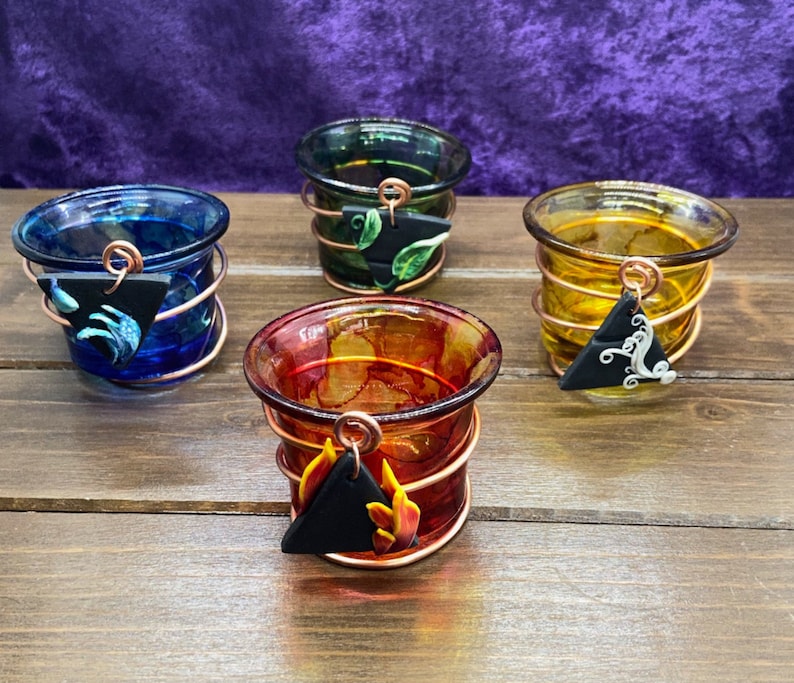 K&ouml;nnte beinhalten: Vier kleine Glas-Kerzenhalter mit Kupferdraht umwickelt. Jeder Halter hat einen schwarzen dreieckigen Anh&auml;nger mit einem anderen Design: ein Blatt, eine Welle, eine Flamme und ein Wirbel.