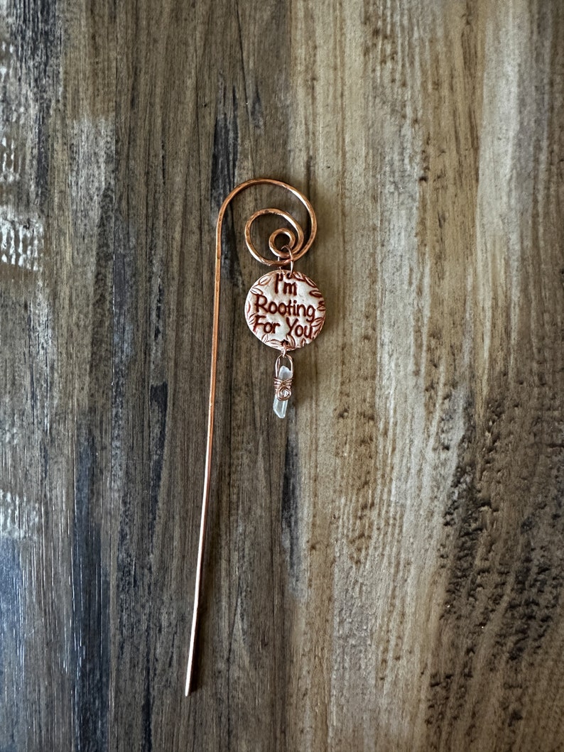 Puede incluir: Un marcap&aacute;ginas de cobre con un amuleto de cer&aacute;mica blanca que dice "Te estoy animando". Un cristal transparente cuelga del amuleto.