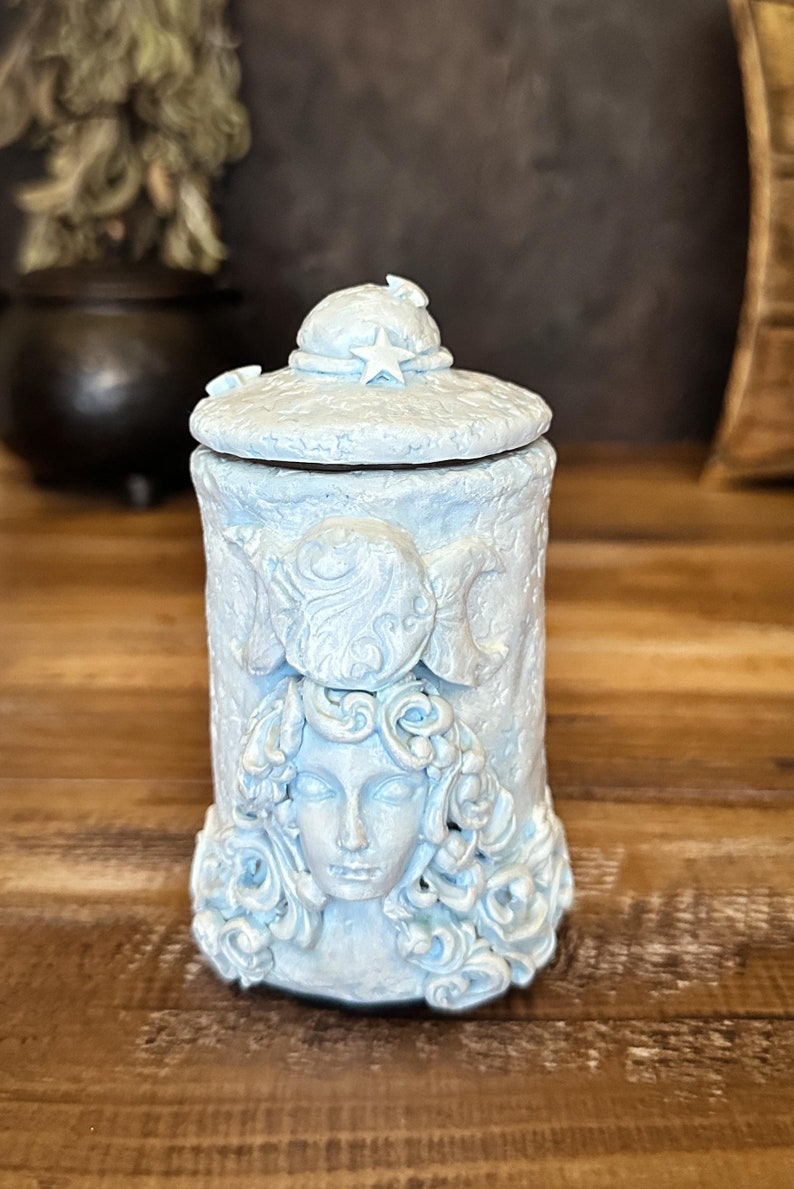 Peut inclure: Un r&eacute;cipient cylindrique bleu clair avec un couvercle amovible orn&eacute; d'une &eacute;toile. La surface ext&eacute;rieure pr&eacute;sente un visage sculpt&eacute; entour&eacute; de cheveux fluides, surmont&eacute; d'un symbole de triple lune. La texture est mate et l&eacute;g&egrave;rement us&eacute;e.
