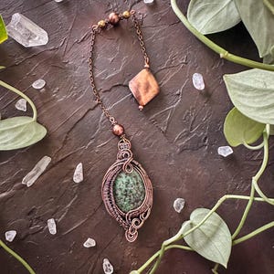 May include: A handcrafted copper pendant necklace featuring a green stone set in a wire-wrapped design. The necklace has a chain with decorative beads and a diamond-shaped charm. The jewelry is displayed on a dark brown surface with greenery and crystals.