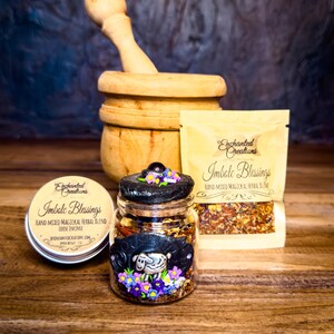 May include: A glass jar of Imbolc Blessings loose incense with a black lid and a sculpted sheep and flower design, accompanied by a matching metal tin and a beige pouch, all labeled Enchanted Creations, set before a wooden mortar and pestle.