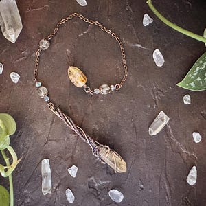 May include: A copper-toned metal chain pendulum featuring a long, pointed, translucent yellow crystal wrapped in intricate copper wire. The chain is accented with smaller clear and yellow crystal beads, creating a decorative hanging ornament.