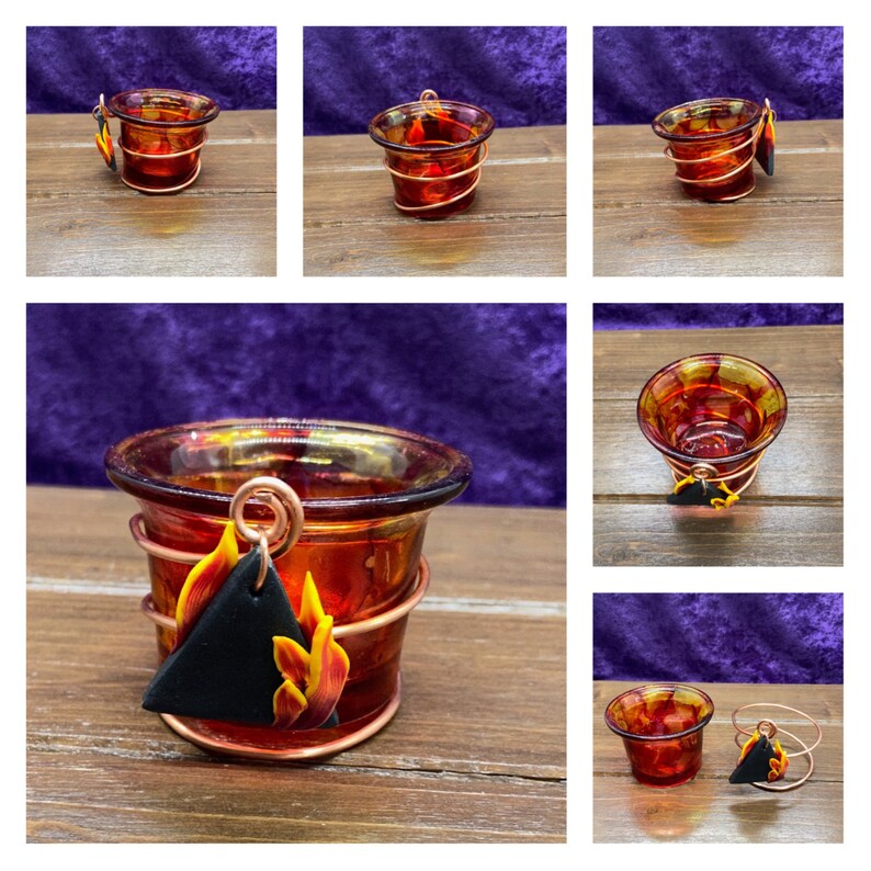 K&ouml;nnte beinhalten: Ein kleiner gl&auml;serner Votivkerzenhalter mit einer Kupferdrahtumwicklung und einem schwarzen und orangefarbenen Ton-Flammenanh&auml;nger. Der Kerzenhalter steht auf einer Holzoberfl&auml;che.