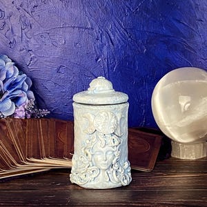 Peut inclure: Un pot cylindrique en c&eacute;ramique bleu clair avec un couvercle, pr&eacute;sentant une sculpture en relief d'un visage entour&eacute; de cheveux tourbillonnants et d'un symbole de triple lune sur le devant. La surface a une finition mate et d&eacute;taill&eacute;e.