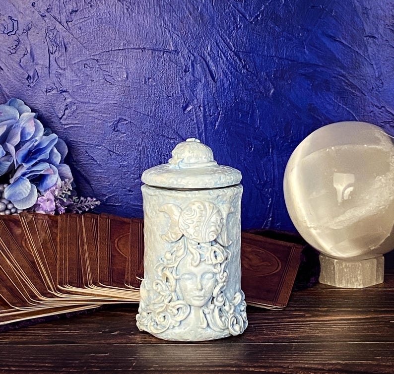 Peut inclure: Un pot cylindrique en c&eacute;ramique bleu clair avec un couvercle, pr&eacute;sentant une sculpture en relief d'un visage entour&eacute; de cheveux tourbillonnants et d'un symbole de triple lune sur le devant. La surface a une finition mate et d&eacute;taill&eacute;e.