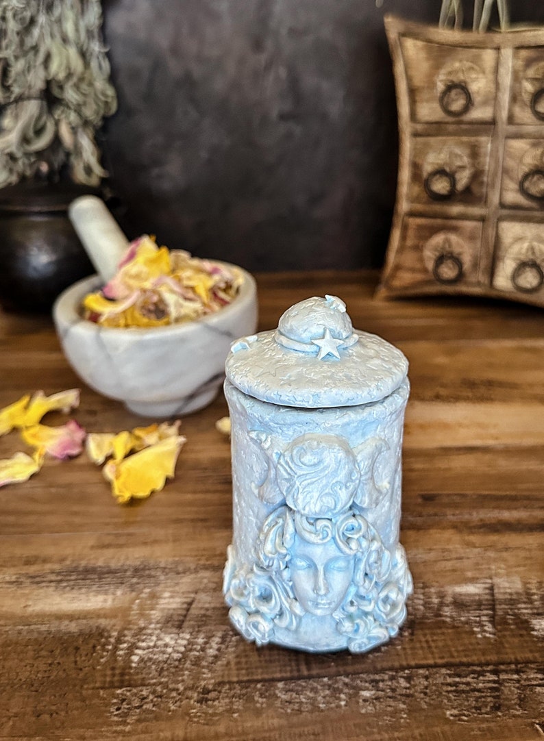 Peut inclure: Un pot cylindrique bleu clair avec un couvercle surmont&eacute; d'une &eacute;toile. La surface ext&eacute;rieure pr&eacute;sente un relief repr&eacute;sentant un visage entour&eacute; de cheveux tourbillonnants et d'un symbole de triple lune. La texture est mate et pierreuse.