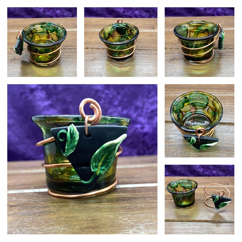 K&ouml;nnte beinhalten: Ein gr&uuml;ner Glas-Votivkerzenhalter mit einem Kupferdrahtk&auml;fig und einem schwarzen und gr&uuml;nen Ton-Blatt-Anh&auml;nger. Der Kerzenhalter steht auf einer Holzoberfl&auml;che.