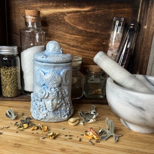 Peut inclure: Un pot en c&eacute;ramique bleu clair avec couvercle, orn&eacute; d'un visage sculpt&eacute; entour&eacute; de cheveux tourbillonnants et d'un symbole de triple lune. Le pot pr&eacute;sente une finition mate textur&eacute;e et est plac&eacute; &agrave; c&ocirc;t&eacute; d'un mortier et pilon en marbre blanc.