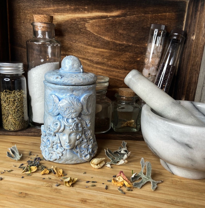 Peut inclure: Un pot en c&eacute;ramique bleu clair avec couvercle, orn&eacute; d'un visage sculpt&eacute; entour&eacute; de cheveux tourbillonnants et d'un symbole de triple lune. Le pot pr&eacute;sente une finition mate textur&eacute;e et est plac&eacute; &agrave; c&ocirc;t&eacute; d'un mortier et pilon en marbre blanc.