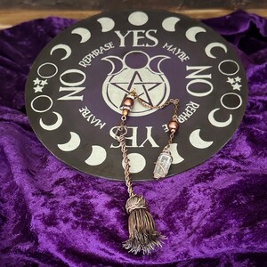 May include: A black circular divination board with a pentagram and the words "YES", "MAYBE", "NO", and "REPHRASE" around the edge. A copper pendulum with a crystal point and a tassel hangs over the board.