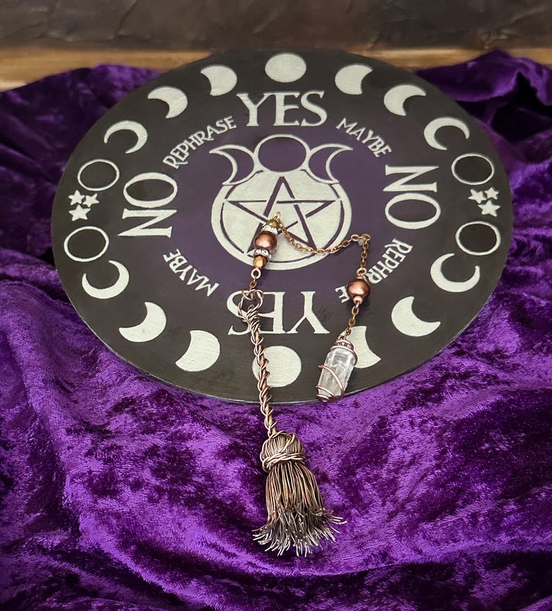 May include: A black circular divination board with a pentagram and the words "YES", "MAYBE", "NO", and "REPHRASE" around the edge. A copper pendulum with a crystal point and a tassel hangs over the board.