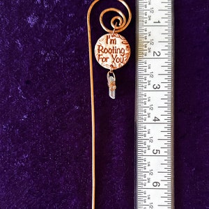 Puede incluir: Un marcap&aacute;ginas dorado con un encanto redondo que dice "I'm Rooting For You". El encanto tiene un fondo blanco con letras marrones y un dise&ntilde;o floral marr&oacute;n. Un peque&ntilde;o cristal transparente cuelga del encanto.