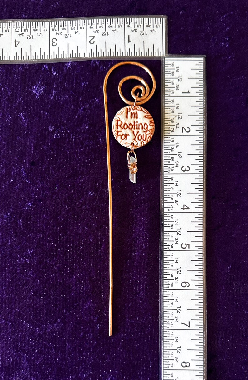 Puede incluir: Un marcap&aacute;ginas dorado con un encanto redondo que dice "I'm Rooting For You". El encanto tiene un fondo blanco con letras marrones y un dise&ntilde;o floral marr&oacute;n. Un peque&ntilde;o cristal transparente cuelga del encanto.