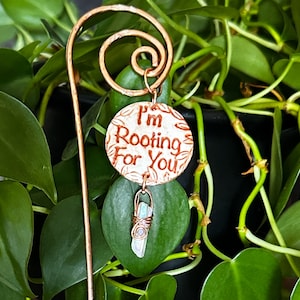 Puede incluir: Marcador de planta de cobre con un amuleto de cer&aacute;mica blanca que dice "Te estoy animando". Un cristal transparente cuelga del amuleto.