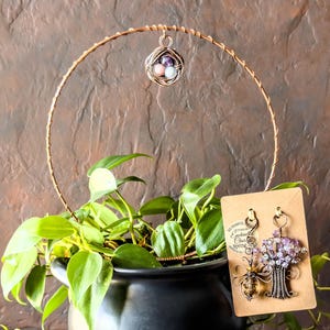 May include: A copper wire circular plant trellis featuring a hanging bird nest charm with three small beads. Beside the plant, a brown card displays two wire-wrapped ornaments: a bee-shaped charm and a tree-shaped charm with purple beads.