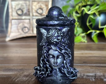 May include: A cylindrical dark purple jar with a matching lid, featuring a sculpted silver-toned face with flowing hair and a moon-shaped emblem on the forehead. The surface is decorated with small star motifs and a textured finish throughout the exterior.