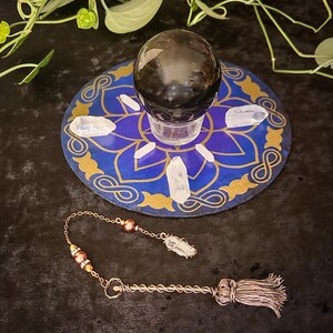 May include: A black obsidian crystal ball sits on a blue and gold mandala with white quartz crystals surrounding it. A copper pendulum with a quartz crystal tip and brown beads hangs below.