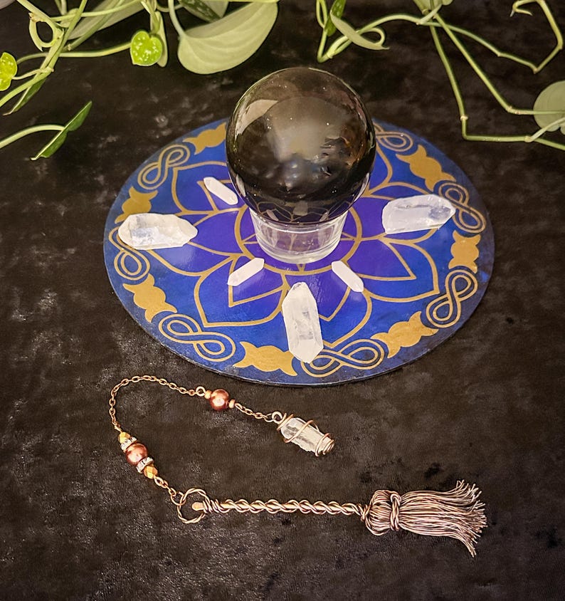 May include: A black obsidian crystal ball sits on a blue and gold mandala with white quartz crystals surrounding it. A copper pendulum with a quartz crystal tip and brown beads hangs below.
