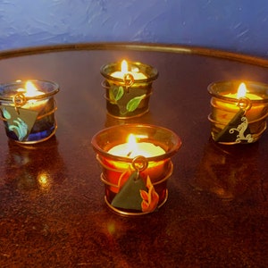 K&ouml;nnte beinhalten: Vier Teelicht-Kerzenhalter mit verschiedenen Glas- und Drahtdesigns. Jeder Halter hat ein kleines dreieckiges Schild mit einem bemalten Design. Die Schilder sind blau mit einem Wellendesign, gr&uuml;n mit einem Blattmuster, braun mit einem Flammenmuster und schwarz mit einem Wirbelmuster.