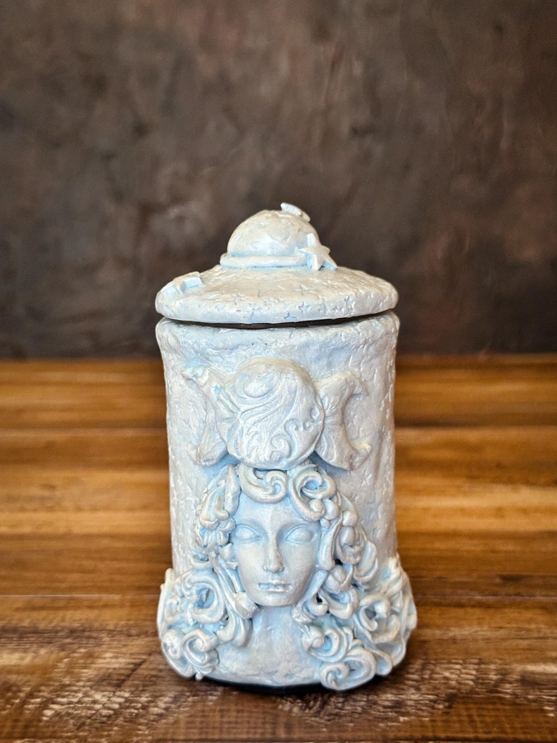 Peut inclure: Un pot cylindrique en c&eacute;ramique bleu clair avec un couvercle assorti. L'ext&eacute;rieur pr&eacute;sente un relief sculpt&eacute; repr&eacute;sentant un visage entour&eacute; de cheveux tourbillonnants, surmont&eacute; d'un symbole de triple lune. Le couvercle est orn&eacute; d'&eacute;toiles.