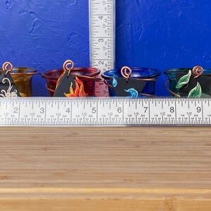 K&ouml;nnte beinhalten: F&uuml;nf kleine Glas-Kerzenhalter mit unterschiedlich gef&auml;rbtem Glas und bemalten Designs. Jeder Halter hat ein kleines schwarzes Schild mit einem anderen Symbol darauf. Die Halter sind in einer Reihe auf einer Holzoberfl&auml;che angeordnet.