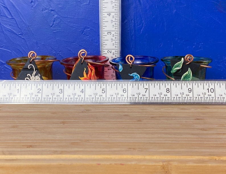 K&ouml;nnte beinhalten: F&uuml;nf kleine Glas-Kerzenhalter mit unterschiedlich gef&auml;rbtem Glas und bemalten Designs. Jeder Halter hat ein kleines schwarzes Schild mit einem anderen Symbol darauf. Die Halter sind in einer Reihe auf einer Holzoberfl&auml;che angeordnet.