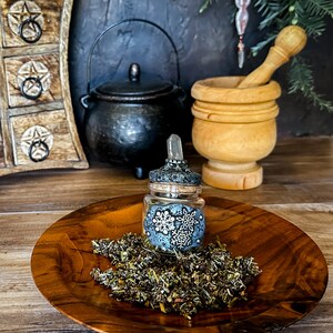 May include: A small glass bottle with a clear glass dropper and a decorative lid with a snowflake design. The bottle is filled with dried herbs and is sitting on a wooden plate.