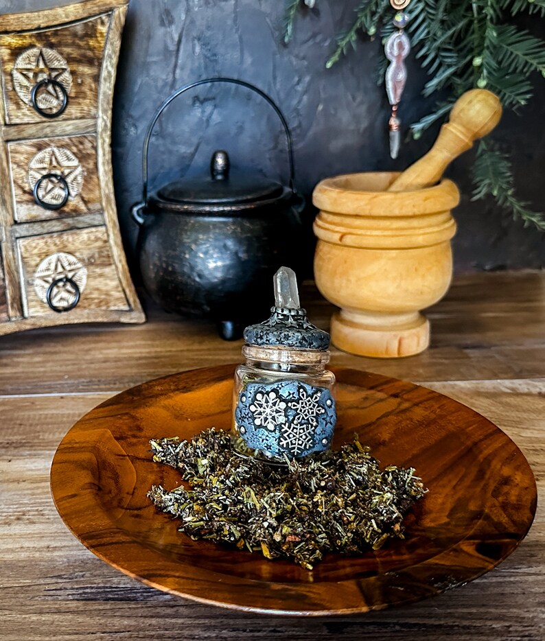 May include: A small glass bottle with a clear glass dropper and a decorative lid with a snowflake design. The bottle is filled with dried herbs and is sitting on a wooden plate.