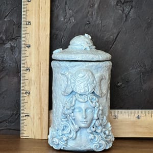 Peut inclure: Un pot cylindrique bleu clair avec couvercle, orn&eacute; d'un relief repr&eacute;sentant un visage aux cheveux boucl&eacute;s et un symbole de triple lune. Une r&egrave;gle en bois plac&eacute;e verticalement et horizontalement indique une hauteur d'environ 11,5 cm.