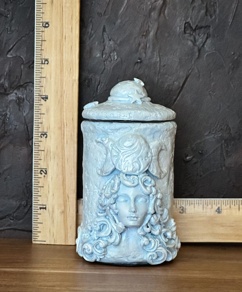 Peut inclure: Un pot cylindrique bleu clair avec couvercle, orn&eacute; d'un relief repr&eacute;sentant un visage aux cheveux boucl&eacute;s et un symbole de triple lune. Une r&egrave;gle en bois plac&eacute;e verticalement et horizontalement indique une hauteur d'environ 11,5 cm.