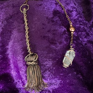 May include: A gold-toned pendulum with a clear quartz crystal and a tassel. The pendulum is hanging from a chain with brown beads and rhinestones.
